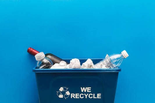 Ein Recyclingbehälter mit der Aufschrift 'Wir recyceln', gefüllt mit verschiedenen Abfällen wie Plastikflaschen und zerknülltem Papier, vor einem blauen Hintergrund.