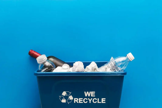 Ein Recyclingbehälter mit der Aufschrift 'Wir recyceln', gefüllt mit verschiedenen Abfällen wie Plastikflaschen und zerknülltem Papier, vor einem blauen Hintergrund.