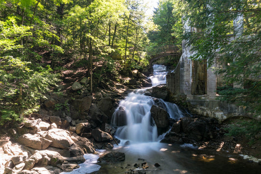 Ein ruhiger Wasserfall, der über Felsen fließt, umgeben von üppigen grünen Bäumen. Rechts befindet sich eine alte Steinstruktur mit einer Brücke über dem Wasserfall, die natürliche Schönheit mit einem Hauch historischer Architektur verbindet.